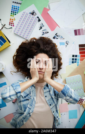 Above view portrait of stressed young woman looking at camera while lying on floor surrounded by art supplies and designer work, copy space Stock Photo