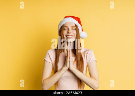 Cheerful caucasian freckled redhead woman in Santa hat making praying gesture, keeping palms together, smiling broadly, isolated over yellow backgroun Stock Photo