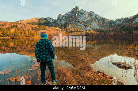 5 years old boy standing at the shore of the Laudachsee, the impressive Katzenstein rising in the background and reflecting in the crystal clear water Stock Photo