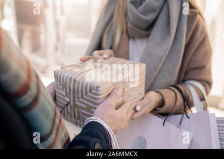 Hands of young man passing wrapped giftbox to his girlfriend with paperbags Stock Photo
