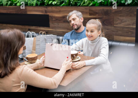 Pretty girl and her mother with paperbag looking at one another by table in cafe Stock Photo