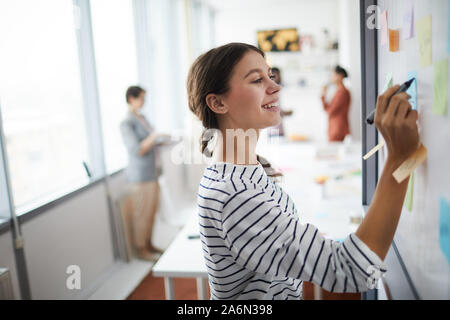 Waist up portrait of smiling young woman writing on whiteboard in office, copy space Stock Photo