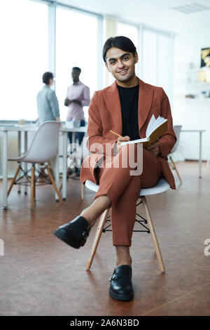 Full length portrait of young Middle-Eastern businessman smiling at camera while sitting on chair in modern office Stock Photo