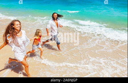 Happy travel family - young father, mother, baby girl hold hands, run together, child jump with fun by water pool along sea surf on tropical beach.