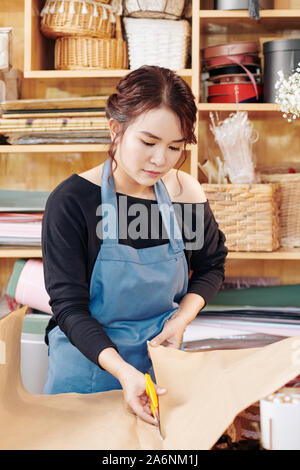 Serious young Asian florist cutting wrapping paper for bouquet or present Stock Photo