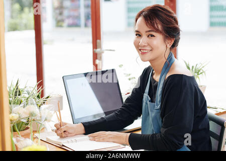 Smiling pretty young florist taking notes in textbook after celling bouquet Stock Photo