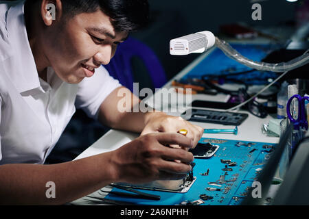 Smiling Asian man repairing smartphone under light of led lamp at his desk Stock Photo