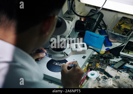 Repairman sitting at desk with broken smartphone parts and looking at logic board through microscope Stock Photo