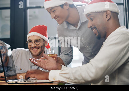 Excited businessman in Santa Claus hat showing finished project on laptop screen to his smiling supportive coworkers Stock Photo