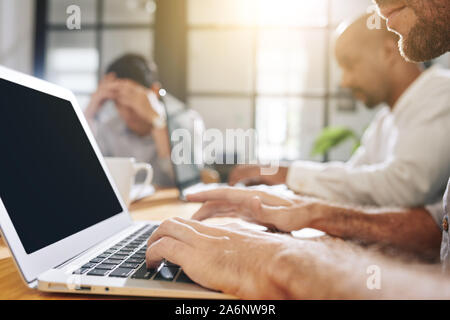 Cropped image of bearded businessman working on laptop whe sitting at big table in meeting room with his colleagues Stock Photo