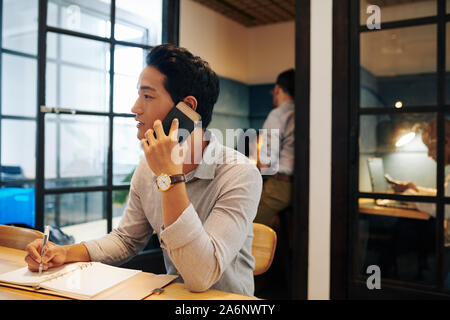 Young Asian businessman sitting at office table, talking on phone with business partner and taking notes Stock Photo