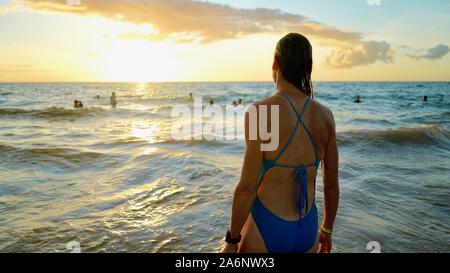 a woman standing by the Pacific Ocean in Hawaii, wearing a blue swimsuit, with wet hair, looking towards setting sign over the horizon Stock Photo
