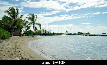 white sandy beach curving around Pacific Ocean shoreline, surrounded by palm trees and cloudy blue sky Stock Photo