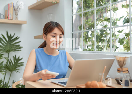 Happy young Asian woman using phone and laptop while working at home behind window. Stock Photo