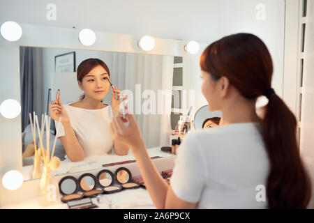 Young Asian woman applying make up (paint her eyelashes) in front of a mirror. Focus on her reflection Stock Photo