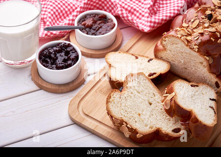 Still life with knitted Christmas cake or sweet pastry with raisins and almonds. Sliced on white wooden board table with bowls with fruit jam, glass o Stock Photo