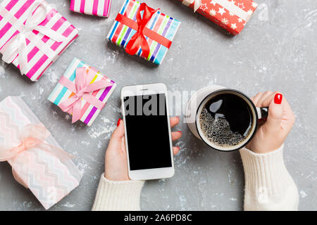 Top view of a woman holding a cup of coffee in one hand and a phone in another hand on cement background. Gift boxes and Christmas decorations. New Ye Stock Photo