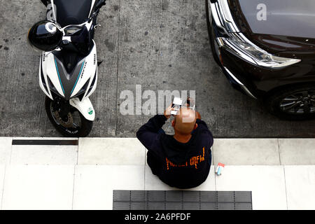 ANTIPOLO CITY, PHILIPPINES – OCTOBER 7, 2019: Man uses smartphone and rests on a sidewalk beside parked motorcycle and car. Stock Photo