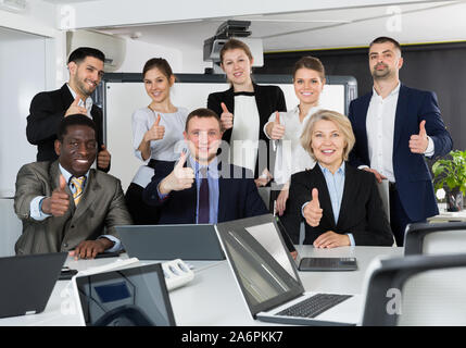 Delighted group of international business people holding thums up in office, enjoying their success Stock Photo
