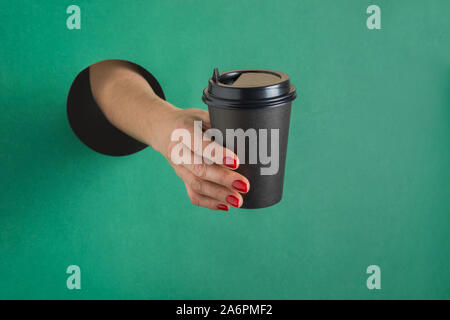 Female hand holding disposable paper black coffee cup through round hole in green paper. Concept Stock Photo