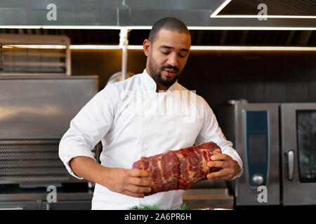 unrecognizable young African chef standing in professional kitchen in restaurant preparing a meal of meat and cheese vegetables. Portrait of man in co Stock Photo