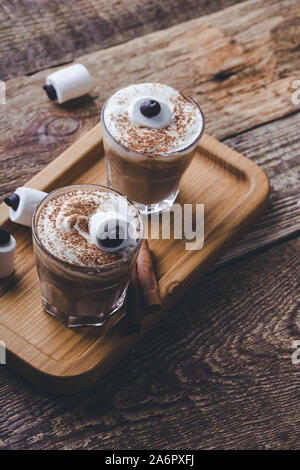 Happy Halloween monster, pumpkin spice latte with whipped cream and big marshmallow eye on top, Halloween dessert in glasses Stock Photo