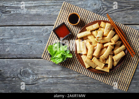 deep-fried crispy bite-sized chinese spring rolls with cabbage, carrots and green peas fillings on a  plate on a rustic wooden table with sauces, view Stock Photo