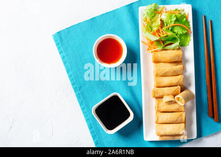 crispy bite-sized Chinese spring rolls with cabbage, carrots and green peas fillings on a white rectangular plate on a wooden table with sauces and sa Stock Photo
