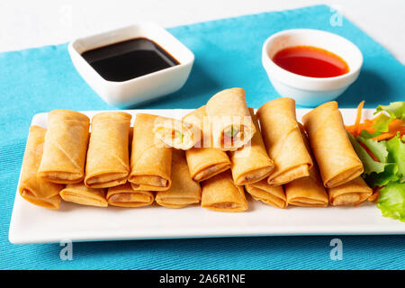 crispy bite-sized Chinese spring rolls with cabbage, carrots and green peas fillings on a white rectangular plate on a wooden table with sauces and sa Stock Photo