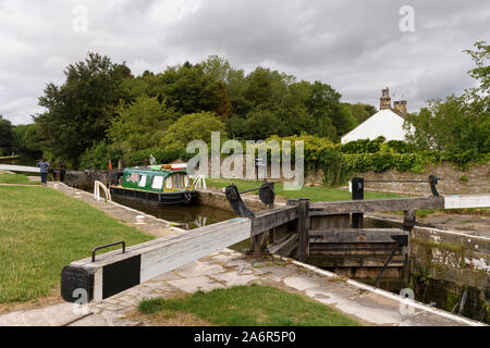 Narrowboat (narrow boat) sailing on scenic rural Leeds Liverpool Canal passing through lock (people by gates) - Gargrave, North Yorkshire, England, UK Stock Photo