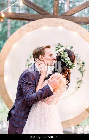 The newlywed stylish couple is hugging and kissing near the wedding arch in the restaurant in rustic style outdoors. Stock Photo