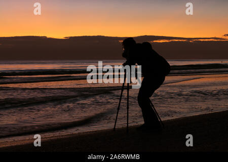 Silhoutte of professional photographer taking pictures with a camera and a tripod in the beach at sunrise. Beautiful sunrise on the background Stock Photo