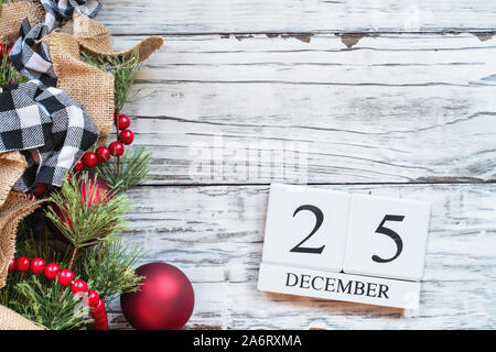 Wood calendar blocks with the date December 25th to mark Christmas Day with ornaments, black and white buffalo check and red bead garland over white r Stock Photo