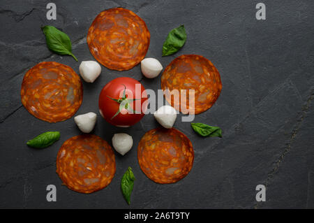 Traditional Italian pizza ingredients mozzarella, salami, tomato and basil leaves composed in a flower shape on black natural stone background. Stock Photo