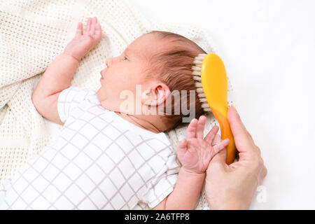 Mother brushing hair of her newborn baby using soft natural bristle hairbrush to stimulate infant’s hair follicles and increase scalp blood flow for h Stock Photo