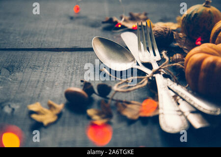 Thanksgiving dinner concept with old cutlery, pumpkins,  dry leaves and orange garland of lights on a blue wooden table. Film grain added Stock Photo