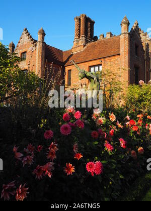 Portrait of Chenies Manor from the west, framed by blue sky, rose bushes and Dahlia varieties.A sculpture reaches up above pink and orange dahlias. Stock Photo