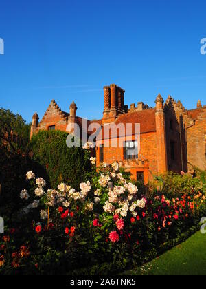 Portrait view of Chenies Manor from the west, framed by blue sky, pastel pink rose bushes and Dahlia varieties, 'Karma Fuschiana, lawn and box edging. Stock Photo