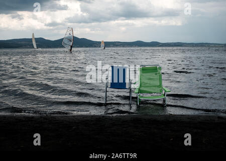 Chairs on the black sand beach of the volcanic lake Bracciano, near Rome, in a cloudy day , with windsurfers in the background Stock Photo
