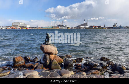 COPENHAGEN, DENMARK - MARCH 16, 2018: the famous Little Mermaid statue in front smoking chimneys of factories in the background.