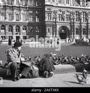 1950s, historical, a French gentleman in a beret and raincoat with his granddaughter sitting feeding the pigeons on the square infront of the Hotel de Ville, Paris, France. Stock Photo