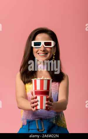 happy young stylish disco girl in 3d glasses holding striped paper bucket with popcorn isolated on pink Stock Photo