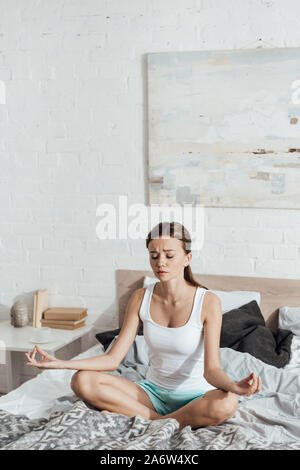 upset young woman sitting on bed in lotus pose with closed eyes Stock Photo