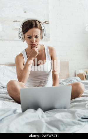 pensive young woman listening music in headphones and using laptop on bed Stock Photo