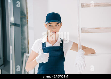 pretty young painter in uniform showing thumb up while looking at camera Stock Photo