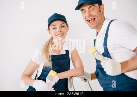 two cheerful painters in uniform holding paintbrushes and smiling at camera Stock Photo