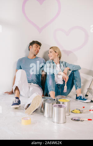happy young couple sitting on floor by white wall under drawn pink hearts Stock Photo