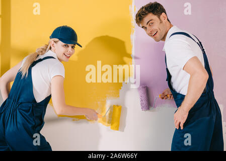 two cheerful painters looking at camera while painting wall in yellow and pink Stock Photo