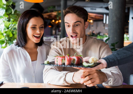 cropped view of waiter holding plate with tasty sushi near surprised couple in sushi bar Stock Photo