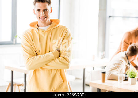 cheerful young businessman smiling at camera while standing with crossed arms in office Stock Photo
